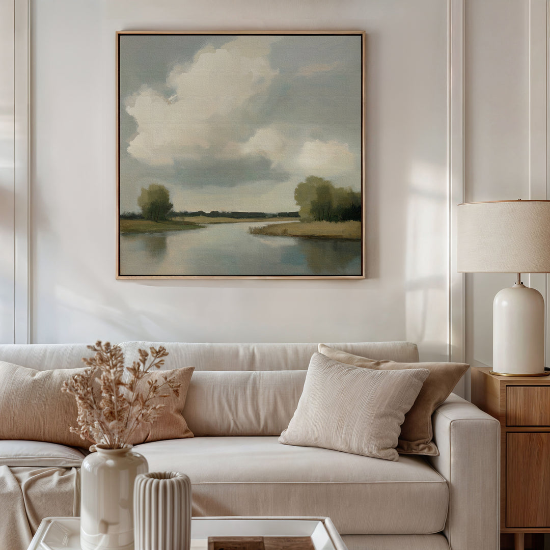 A cozy living room with a beige sofa, neutral pillows, a ceramic vase with dried flowers on the coffee table, a side table with a lamp, and the Calm Passage Canvas Art—a large painting of clouds and water—hanging on the wall.