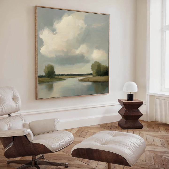 A modern living room featuring a white leather lounge chair and ottoman, a small wooden side table with a white lamp, and the Calm Passage Canvas Art—a large minimalist river-and-cloud painting—creates an inviting, tranquil space.
