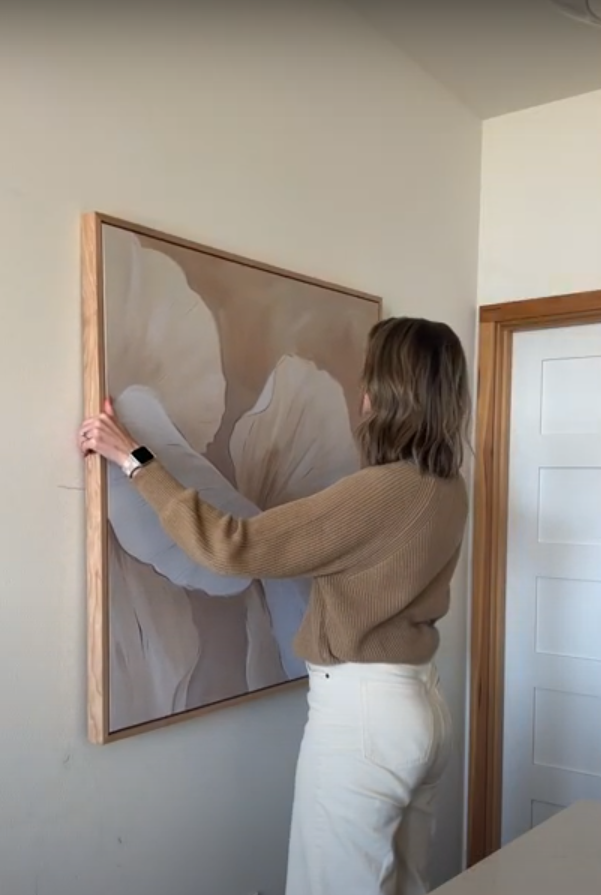 A woman in a brown sweater and white pants hangs a large abstract floral painting with neutral tones on a beige wall near a white door.