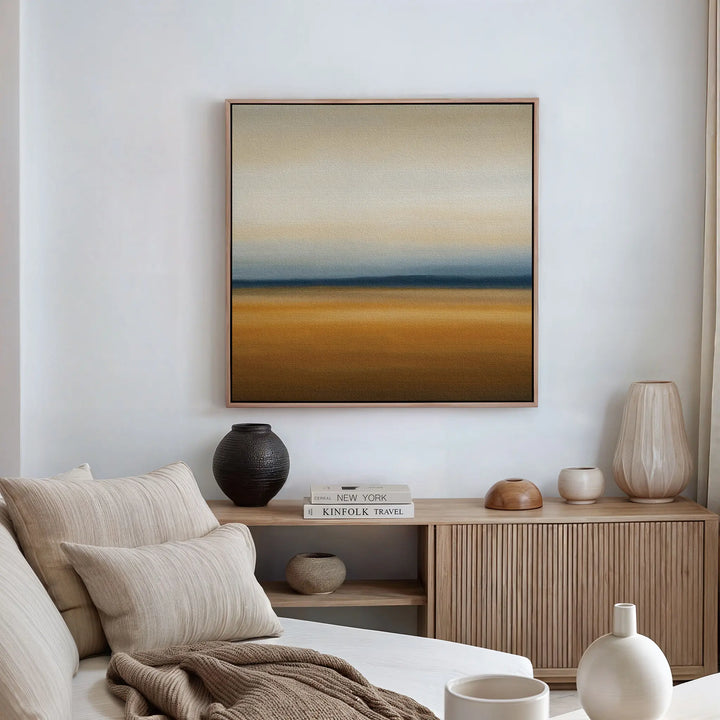 A minimal, modern living room with neutral tones features a beige sofa, earth-toned pillows, a wooden sideboard with decor, and the Caramel Plains Canvas Art displaying abstract horizontal bands of beige, blue, and brown on the wall.