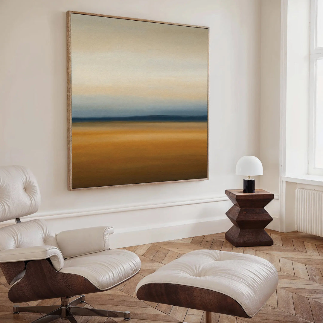 A modern living room with a white lounge chair and ottoman, a geometric wooden side table with a white lamp, and the Caramel Plains Canvas Art in earthy tones creating a bold focal point on the wall.