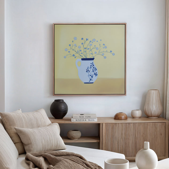 A cozy living room with a beige sofa, neutral cushions, wood sideboard, books, decor items, and the Cerulean Petals Canvas Art—showcasing blue florals in a vase on a yellow background—hanging on the wall.