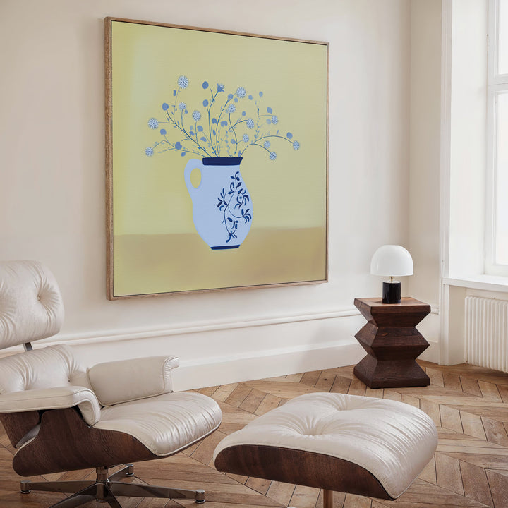 A modern living room with a white leather lounge chair, ottoman, wooden side table with a white lamp, and Cerulean Petals Canvas Art—blue floral artwork on a pale yellow background—displayed above the seating area.