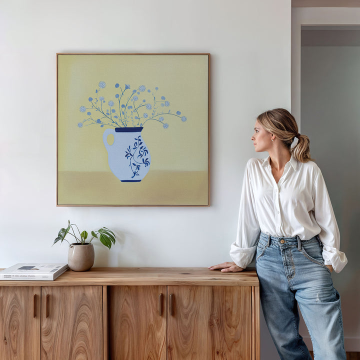 A woman in a white blouse and jeans stands by a wooden cabinet, admiring the Cerulean Petals Canvas Art—a blue floral painting—on a light yellow wall, accented by a small potted plant and books for minimalist decor.