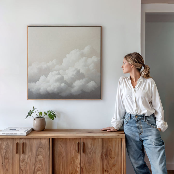 A woman in a white blouse and jeans stands by a wooden cabinet, admiring Cloud Reverie Canvas Art—a minimalist framed painting of fluffy clouds. A potted plant and book on the cabinet complete the calming bedroom decor.