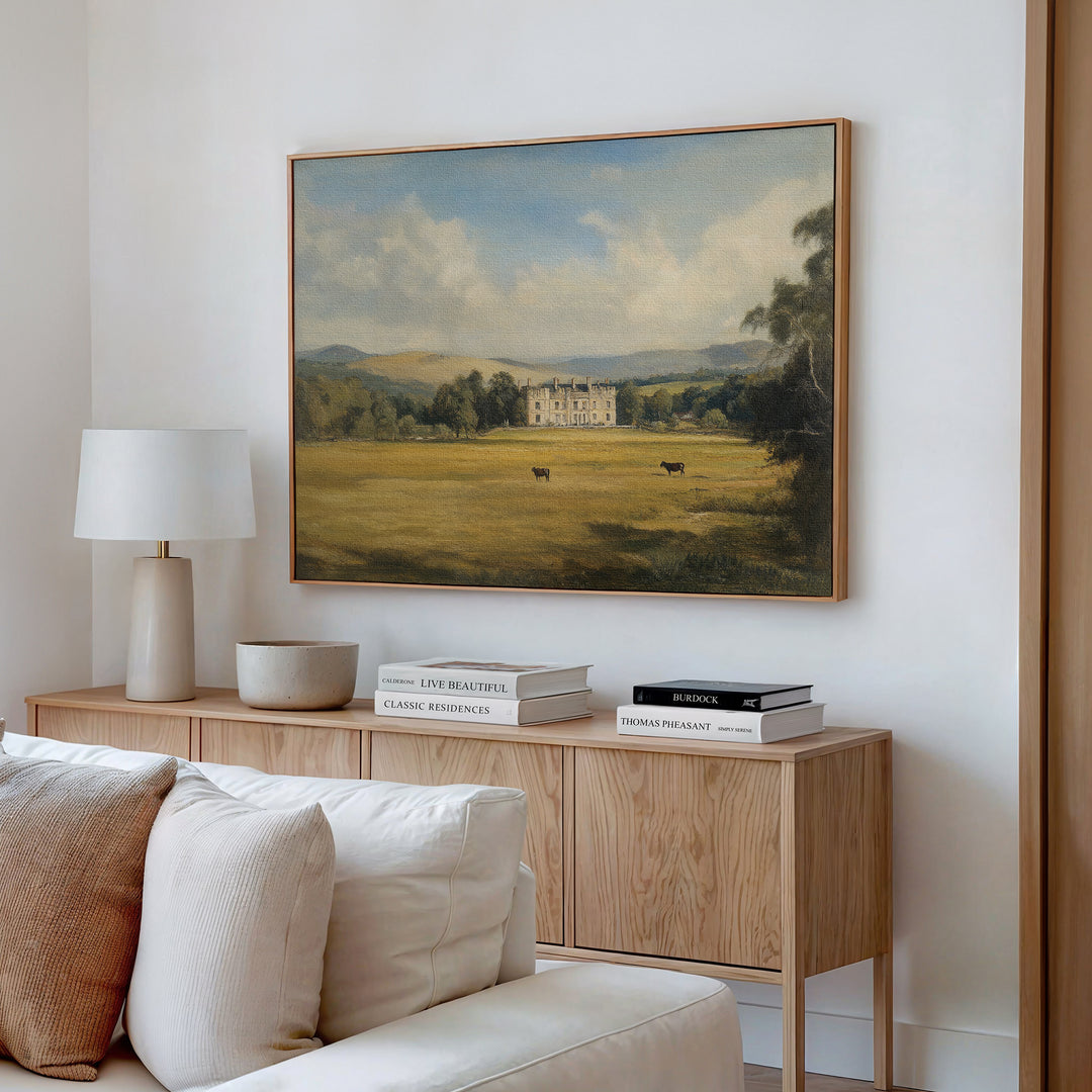 A cozy living room with a light wood sideboard, neutral decor, and a white table lamp. Above the sideboard hangs the Countryside Dream Canvas Art, depicting a manor house in a green field with cows and distant hills.