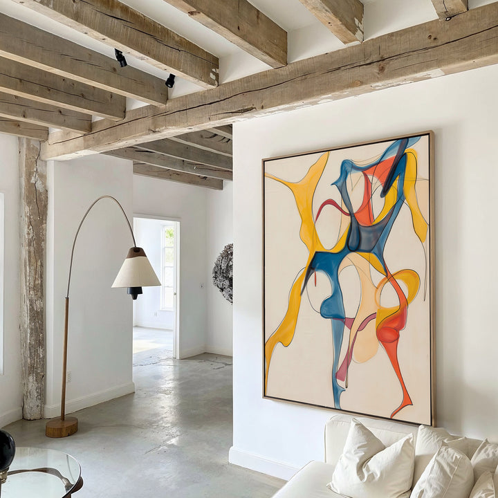 A modern living room features exposed wooden ceiling beams, a curved floor lamp, white walls, and the Dance Of Unity Abstract Canvas Art in blue, yellow, and red tones above a white sofa for a chic decor.