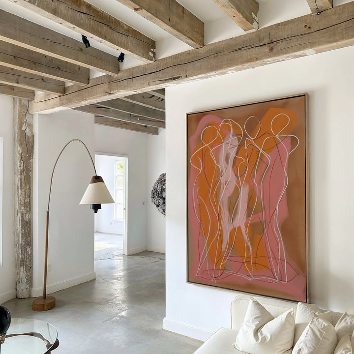 A modern living room with exposed wooden beams, a white sofa, a curved floor lamp, and sunlight streaming in. The wall displays the Dancing Silhouettes Abstract Canvas Art with overlapping orange and pink figures.
