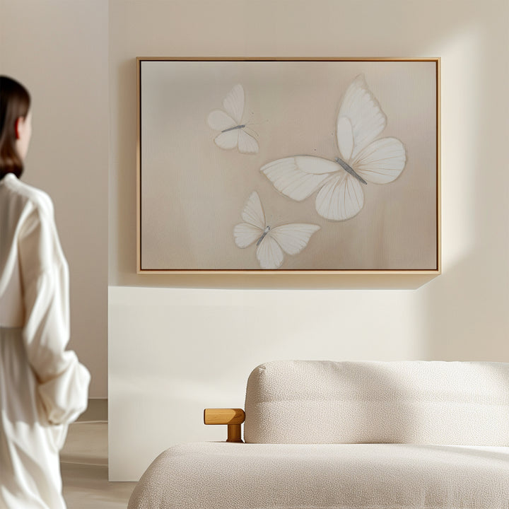 A person admires Delicate Whites Canvas Art—three white butterflies framed—on a beige wall above a light sofa, adding elegant neutral decor to this softly lit, Nordic Park-inspired room.