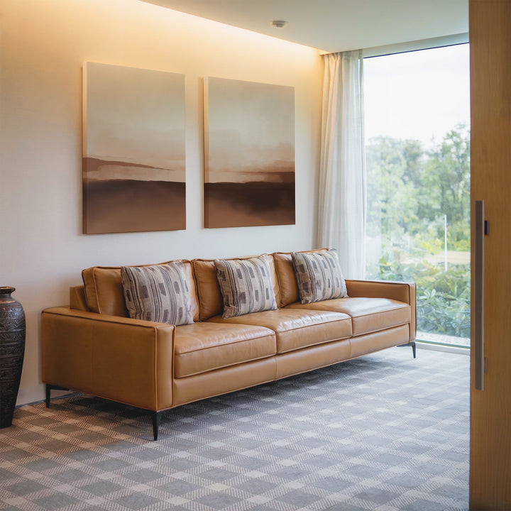 A modern living room featuring a tan leather sofa, four patterned cushions, neutral decor, Desert Landscape Set of 2 minimalist prints on the wall, a large window with sheer curtains, and a ceramic vase on a patterned carpet.