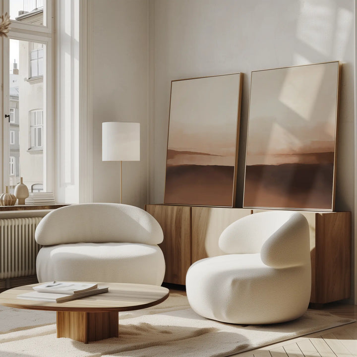 A modern living room with neutral interiors features two white rounded armchairs, a low wooden coffee table, a floor lamp, and the Desert Landscape Set of 2 minimalist prints in earth tones on a light wood sideboard by tall windows.