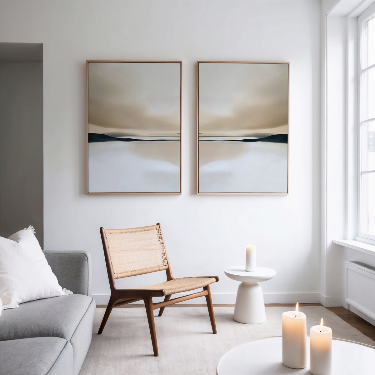 A minimalist living room with a wooden chair, small white table, lit candles, a gray sofa with a white pillow, and the Desert Quietude Set of 2 paintings above. Natural light streams through a large window, highlighting the modern decor.