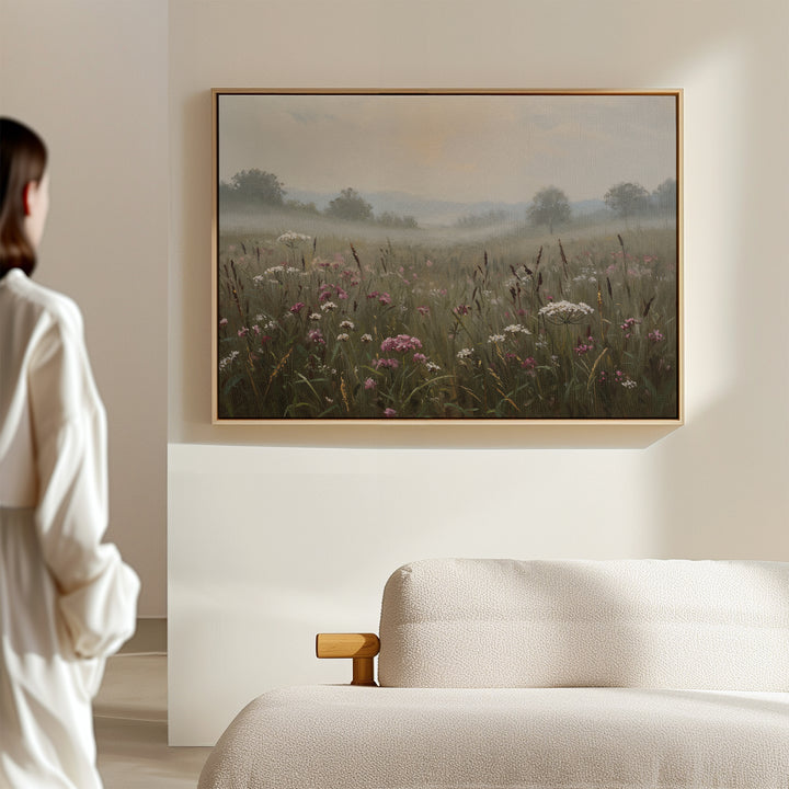 A woman admires Distant Fields Canvas Art—a nature-inspired, large print of a misty wildflower meadow—hanging above a light-colored sofa on a beige wall in a softly lit room.