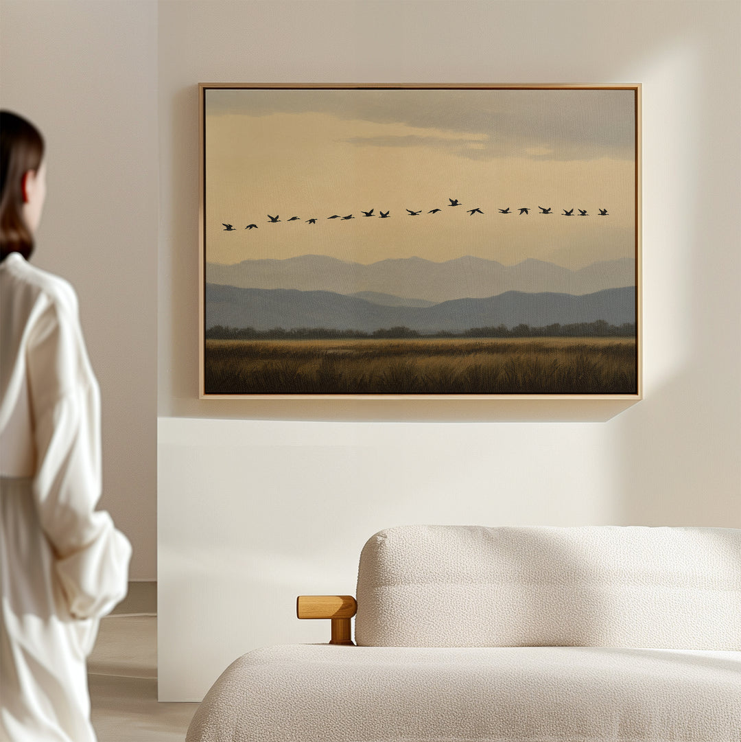 A woman stands in a softly lit room, admiring the Distant Flight Canvas Art—birds soaring above mountains—hung on a light wall over a modern beige sofa, infusing Nordic Park style into the decor.