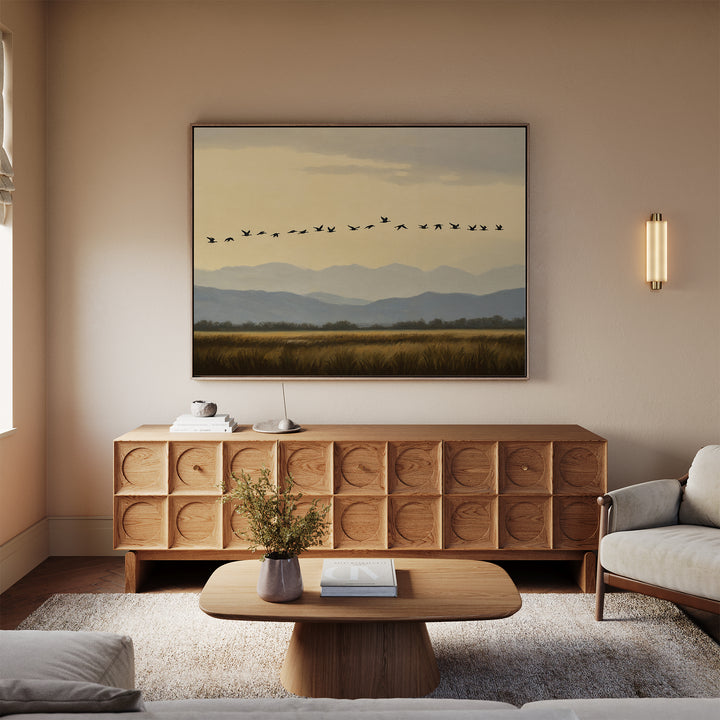 A cozy living room with neutral tones includes a wooden sideboard, low coffee table with a vase of flowers, sofa, and a striking Distant Flight Canvas Art print depicting birds flying over mountains and fields.