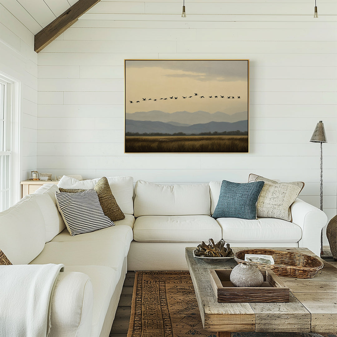 A bright, cozy living room features a white sectional sofa, assorted pillows, a wooden coffee table, rustic decor, and the Distant Flight Canvas Art showcasing birds soaring over mountains on a white shiplap wall.