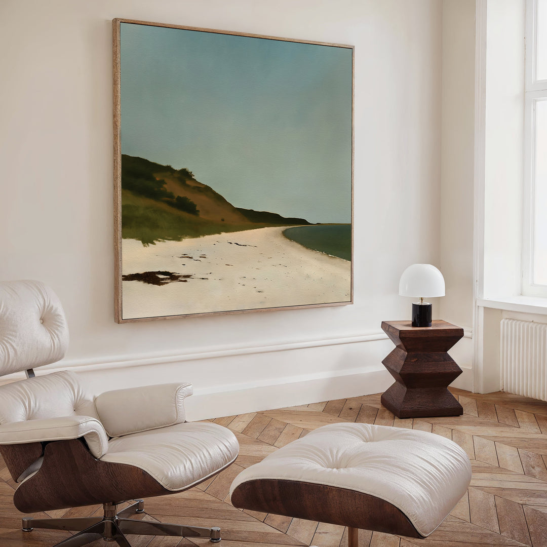 A modern living room with a white lounge chair and ottoman, a wooden side table with a small white lamp, and large wall art—the Drift Shore Canvas Art—displayed on the wall.