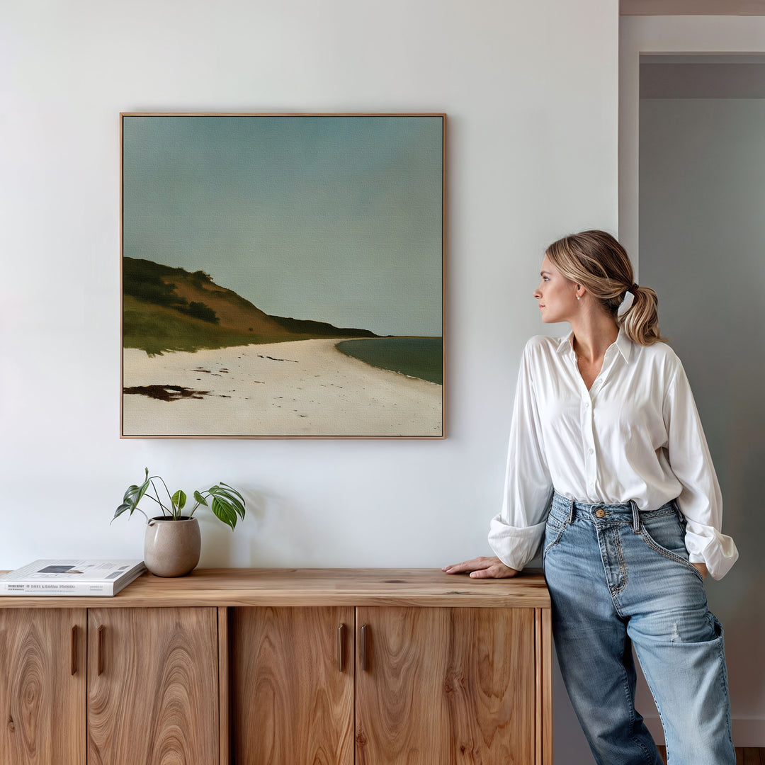 A woman in a white blouse and jeans leans on a wooden sideboard, admiring the Drift Shore Canvas Art—a large print of a sandy beach. A small plant and books on the sideboard add minimalist decor to highlight this coastal wall art.