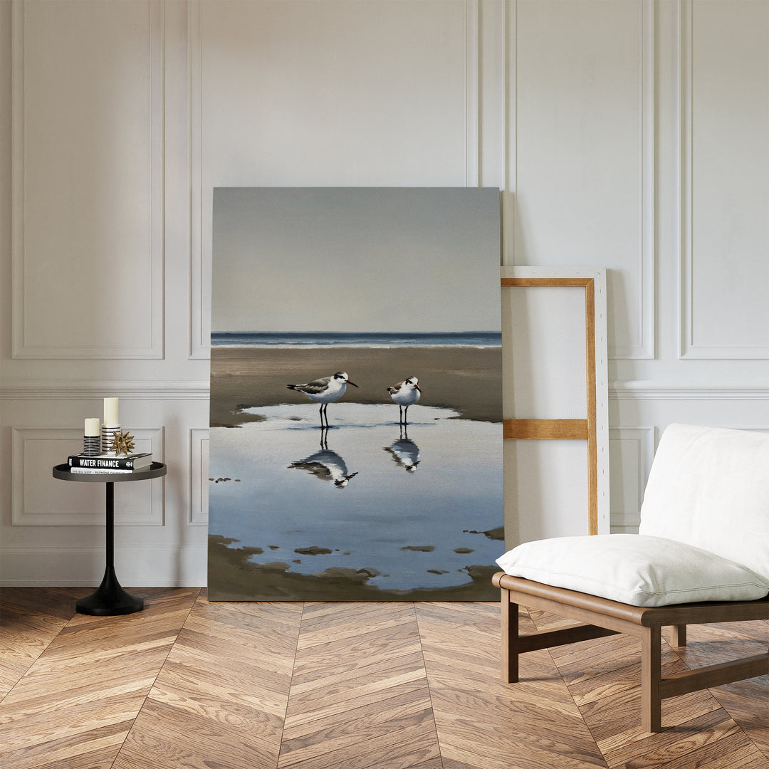 The Drifted Calm Canvas Art, a minimalist wall piece depicting two shorebirds on wet sand with reflections, is displayed against a white paneled wall in a modern interior with a white chair, wood floor, and a round table holding candles and books.