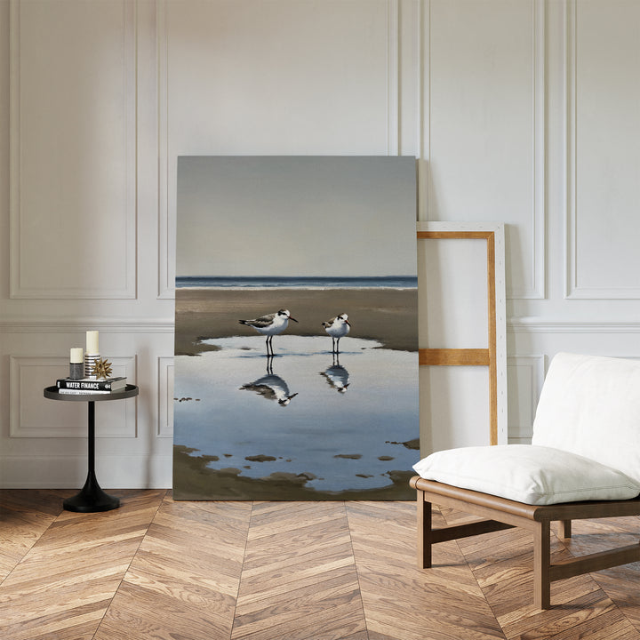 The Drifted Calm Canvas Art, a minimalist wall piece depicting two shorebirds on wet sand with reflections, is displayed against a white paneled wall in a modern interior with a white chair, wood floor, and a round table holding candles and books.