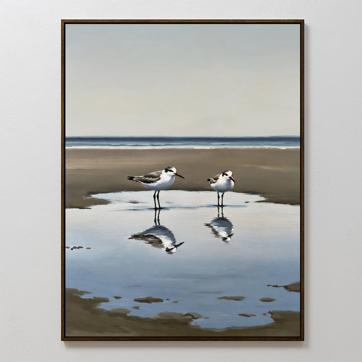 Drifted Calm Canvas Art: A framed painting of two shorebirds wading in a shallow pool on a sandy beach, with their reflections and a peaceful ocean backdrop—perfect minimalist wall art for coastal interiors.