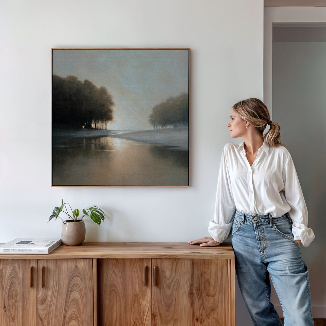 A woman in a white blouse and jeans leans on a wooden cabinet, admiring the Ember Shore Canvas Art above. A plant and books enhance the serene living room decor, highlighting the minimalist charm of the space.