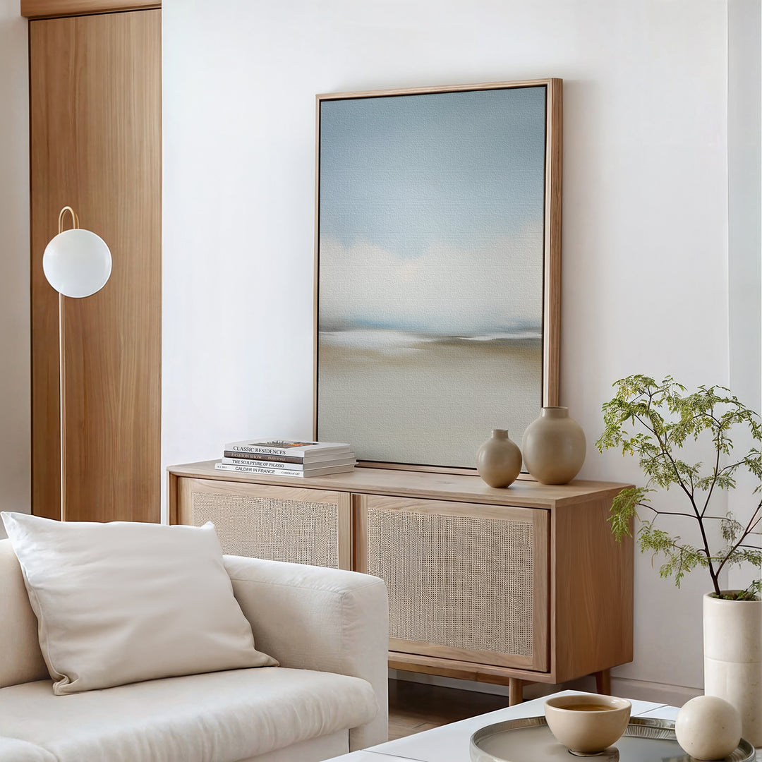 A minimalist living room with a beige sofa, light wood sideboard, neutral vases, stacked books, a potted plant, and a round white pendant lamp features the Faded Silence Canvas Art on white walls.