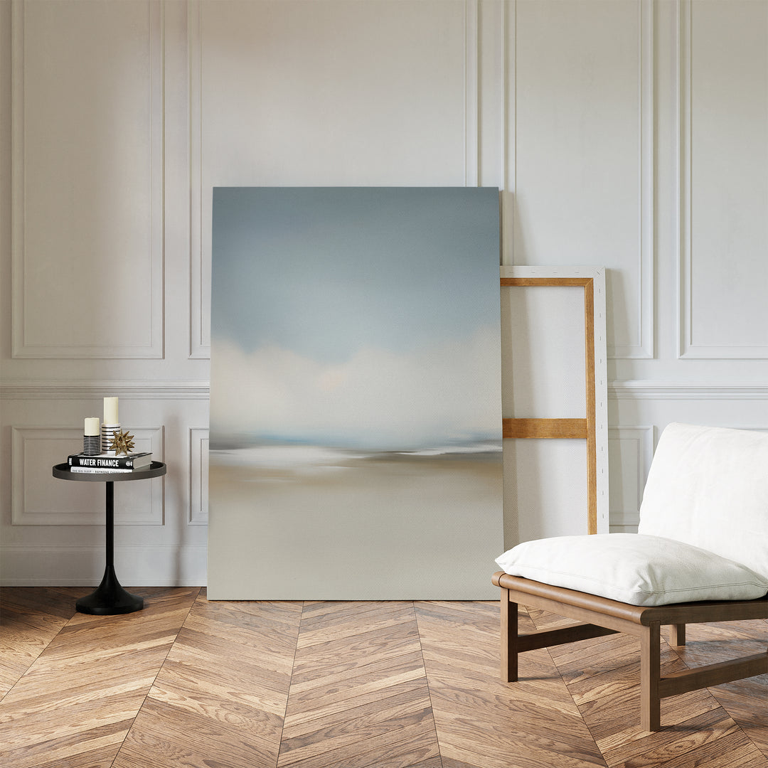 The Faded Silence Canvas Art, featuring minimal softly blurred tones, leans against a white paneled wall beside a cushioned wooden chair and small black table with candles and books atop a herringbone wood floor.