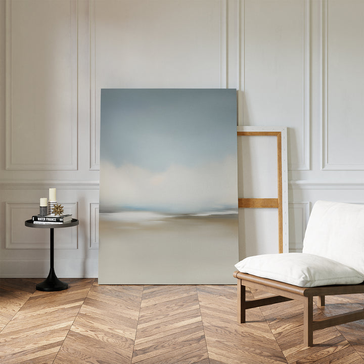 The Faded Silence Canvas Art, featuring minimal softly blurred tones, leans against a white paneled wall beside a cushioned wooden chair and small black table with candles and books atop a herringbone wood floor.