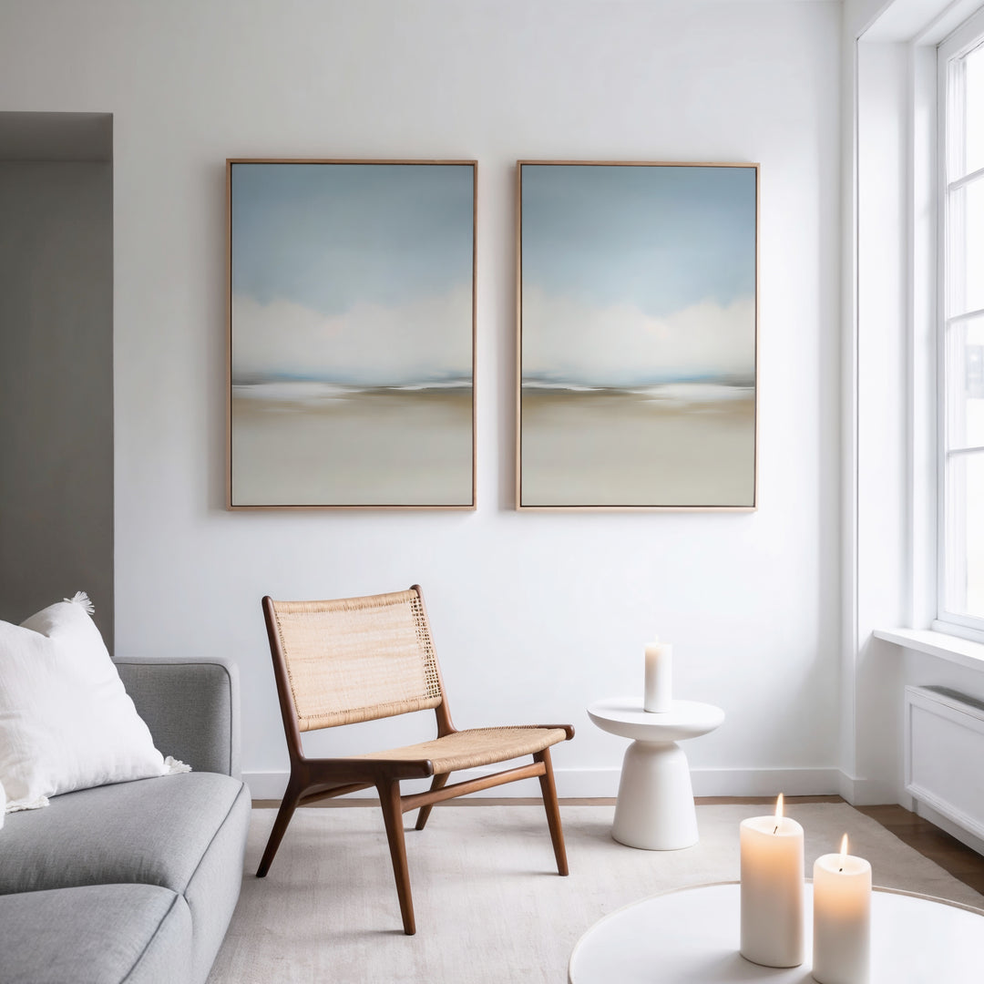 A minimal, modern living room with a gray sofa, woven chair, small white side table, and lit candles. The Faded Silence Set of 2 abstract wall art hangs on a white wall beside a large window, enhancing the serene atmosphere.