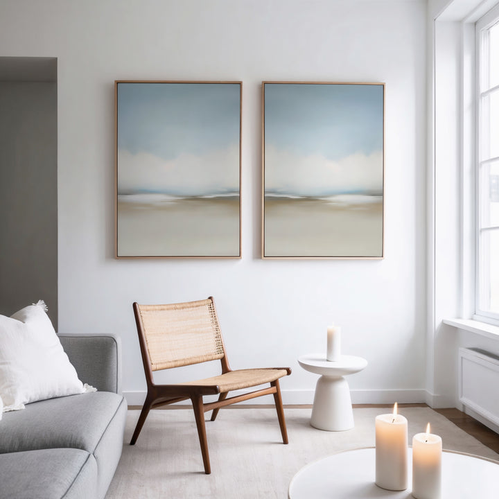 A minimal, modern living room with a gray sofa, woven chair, small white side table, and lit candles. The Faded Silence Set of 2 abstract wall art hangs on a white wall beside a large window, enhancing the serene atmosphere.