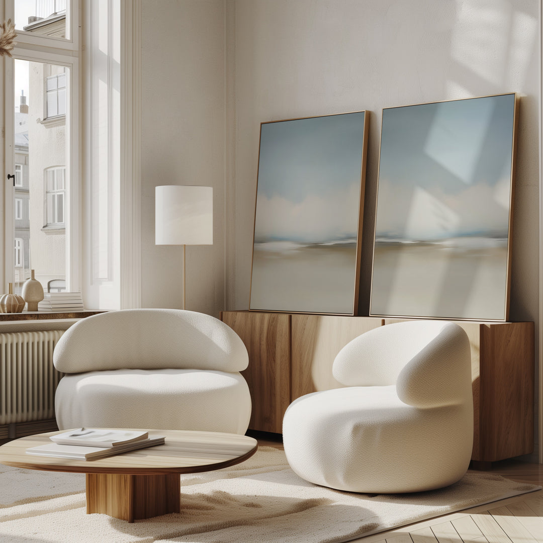 A modern living room features the Faded Silence Set of 2 white rounded armchairs, a low wooden coffee table, a floor lamp, minimalist wall art, and a wooden sideboard by large sunny windows.