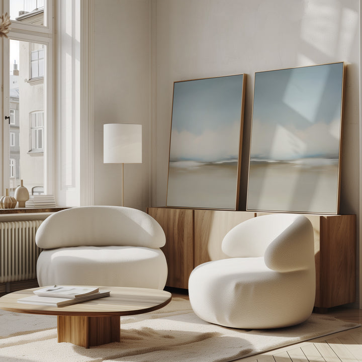 A modern living room features the Faded Silence Set of 2 white rounded armchairs, a low wooden coffee table, a floor lamp, minimalist wall art, and a wooden sideboard by large sunny windows.