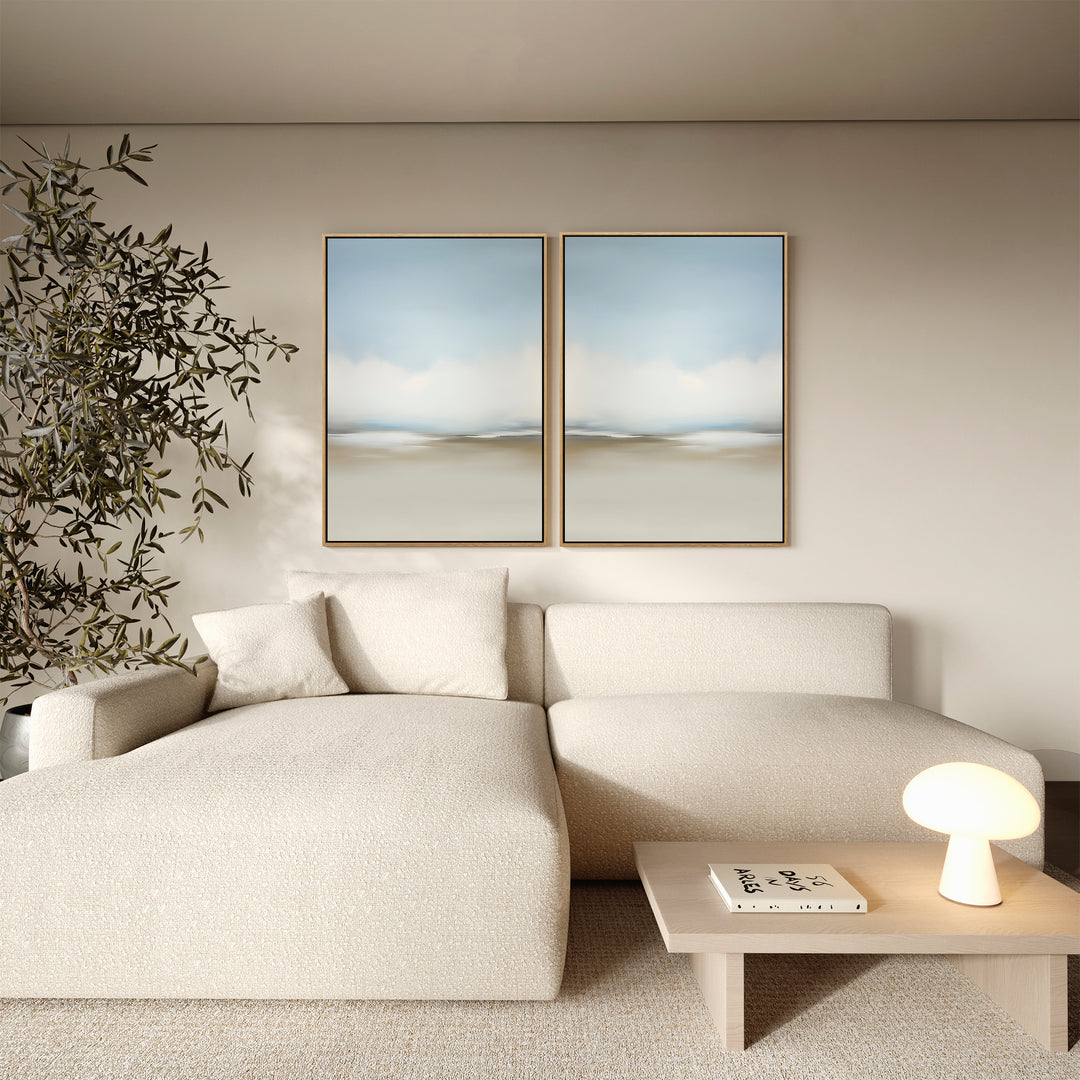 A modern living room features a cream sectional sofa, a small wooden table with a lamp and book, a leafy plant, and minimalist wall art—Faded Silence Set of 2 abstract landscape paintings—displayed above the sofa.
