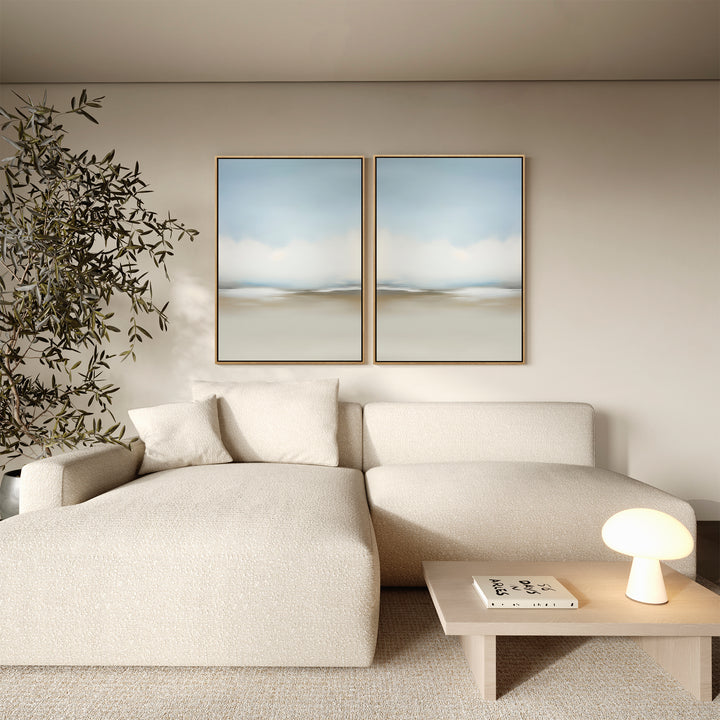 A modern living room features a cream sectional sofa, a small wooden table with a lamp and book, a leafy plant, and minimalist wall art—Faded Silence Set of 2 abstract landscape paintings—displayed above the sofa.