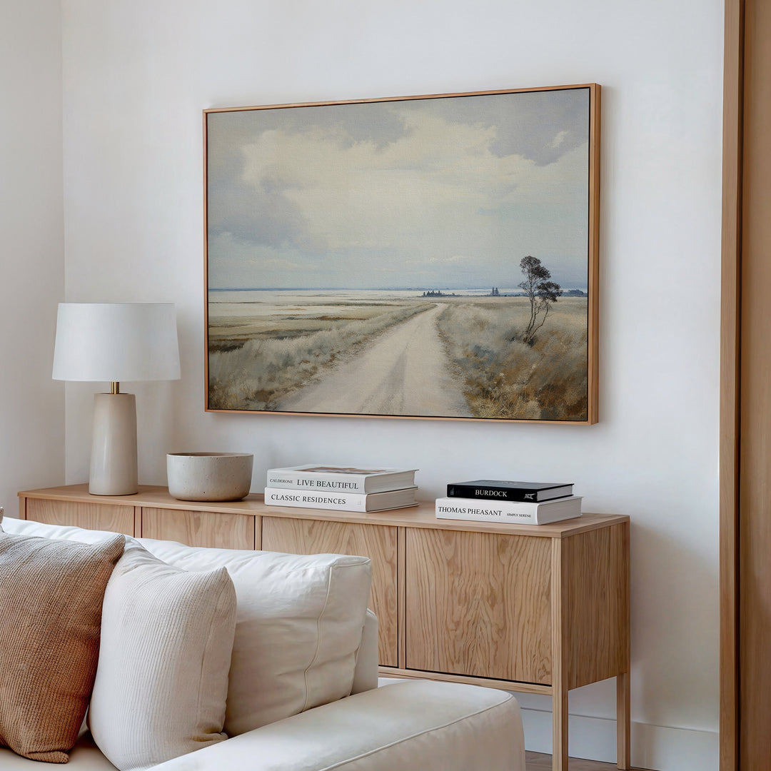 A cozy living room features a wooden sideboard with books, a lamp, and a bowl. Above it hangs the Far Light Canvas Art—minimalist wall art of a rural dirt road with fields and cloudy sky. A beige sofa sits in front.