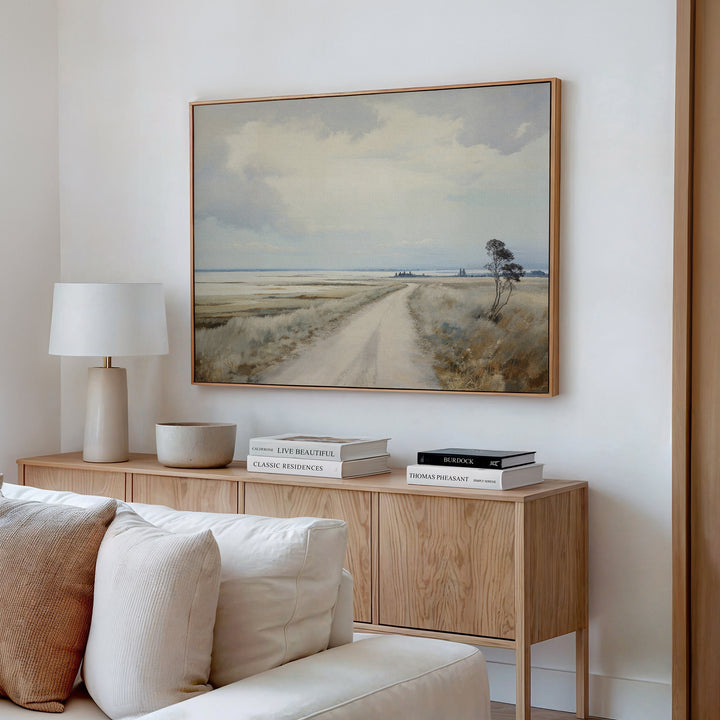 A cozy living room features a wooden sideboard with books, a lamp, and a bowl. Above it hangs the Far Light Canvas Art—minimalist wall art of a rural dirt road with fields and cloudy sky. A beige sofa sits in front.