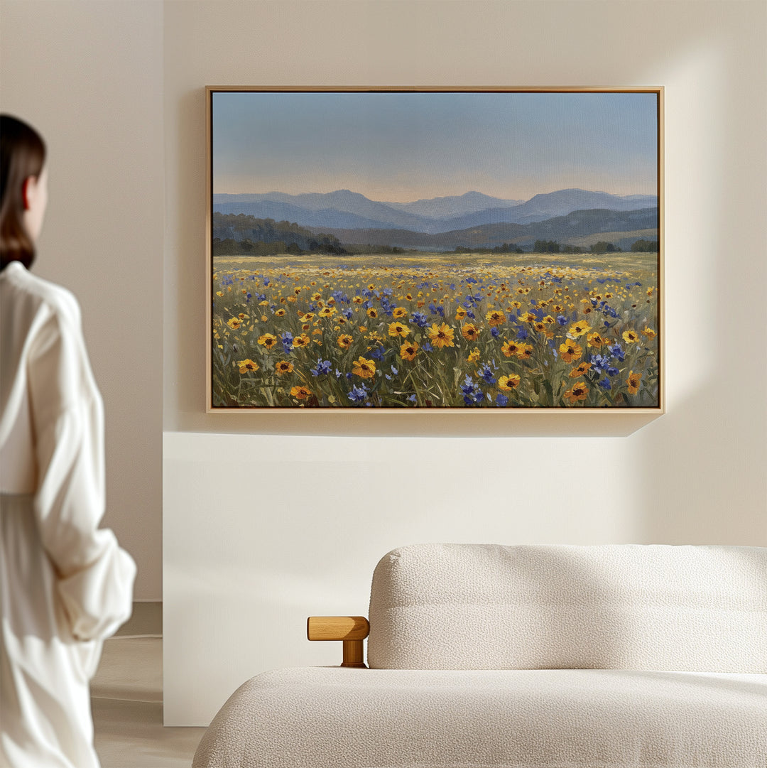 A person admires the Fields of Color Canvas Art, featuring a vibrant wildflower field with yellow and blue flowers, mountains, and a cloudy sky, displayed above a beige sofa as modern home decor.