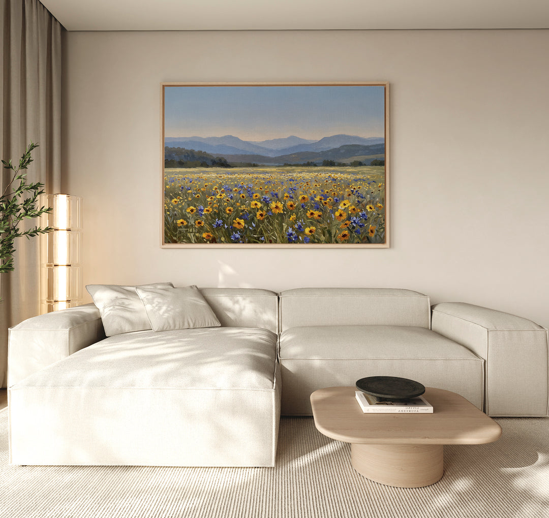 A modern living room with a light beige sectional sofa, a wooden coffee table, and the Fields of Color Canvas Art depicting a wildflower field with mountains hanging on the neutral-colored wall.