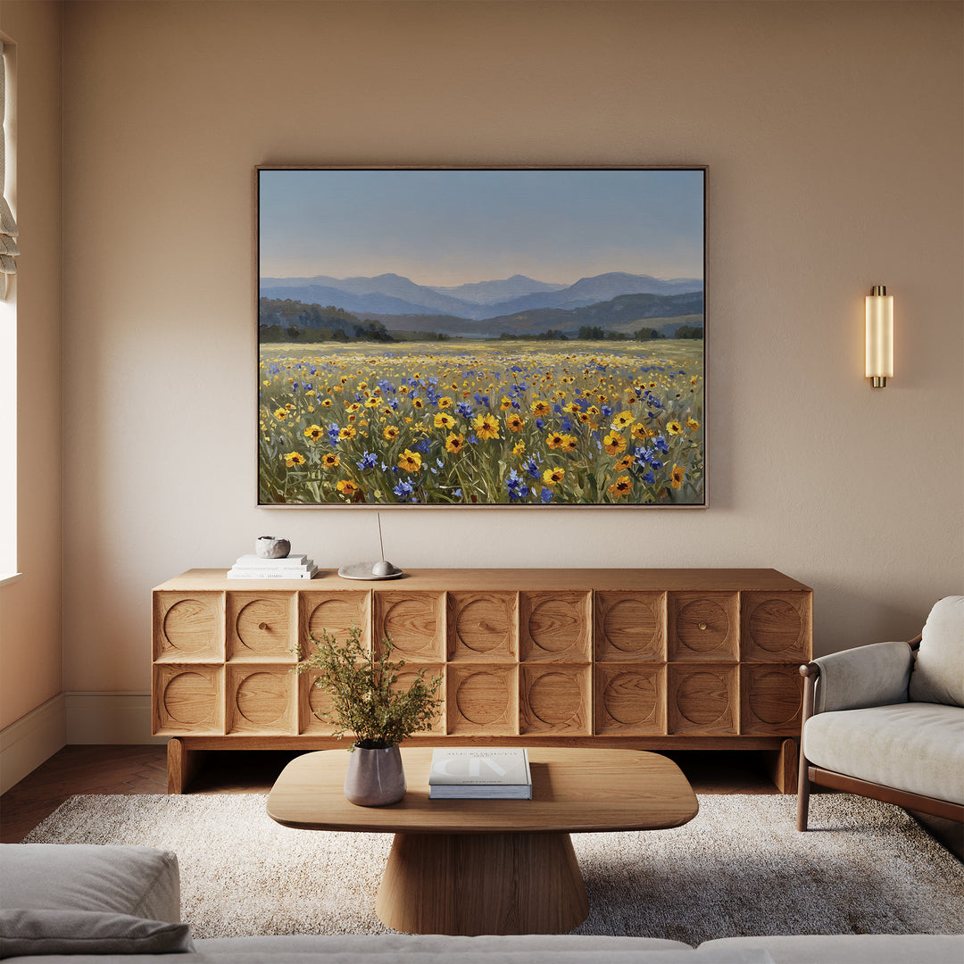 A cozy living room with a wooden coffee table, beige sofa, patterned sideboard, and the Fields of Color Canvas Art featuring wildflower fields and mountains. A vase with flowers on the table completes the modern decor.