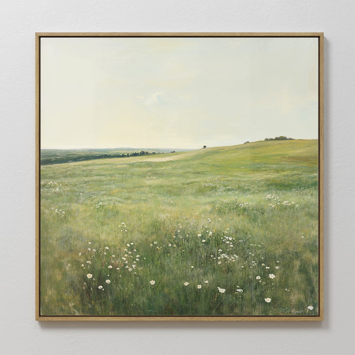 The Floral Meadow Canvas Art, featuring a sunlit grassy meadow with white wildflowers, rolling hills, and a distant tree beneath a pale sky, hangs on a white wall—an ideal touch for living room decor.