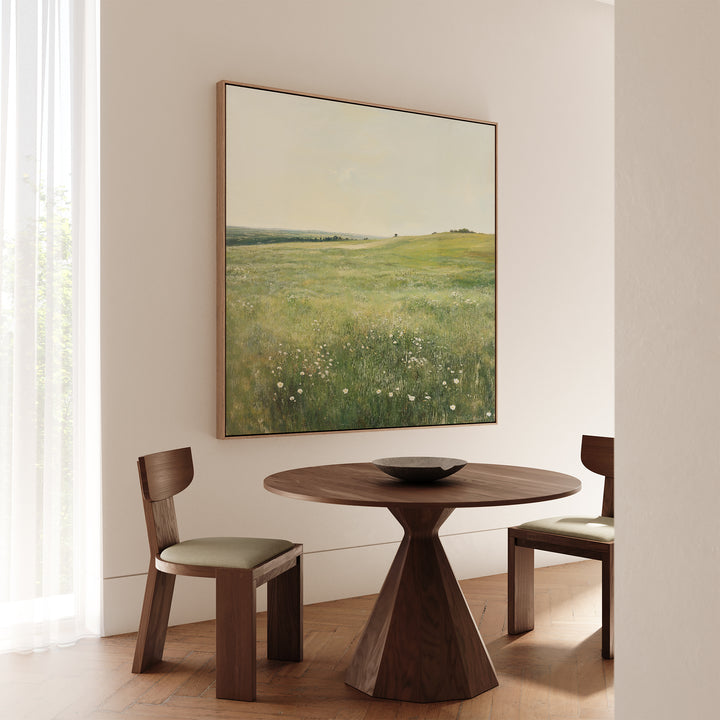 A minimal dining area features a round wooden table, two cushioned chairs, and the Floral Meadow Canvas Art on a white wall. Sunlight through sheer curtains creates a thoughtful living space for decor enthusiasts.
