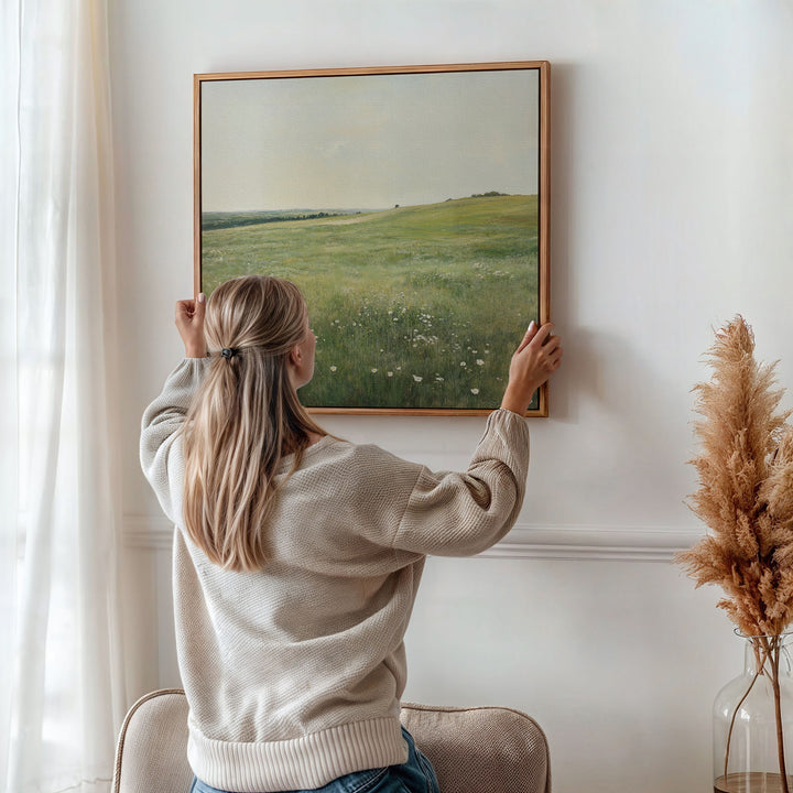 A woman with blonde hair in a low ponytail hangs the Floral Meadow Canvas Art, featuring a green field, on a white wall behind a beige chair. Pampas grass in a vase and sunlight through sheer curtains complete the scene.