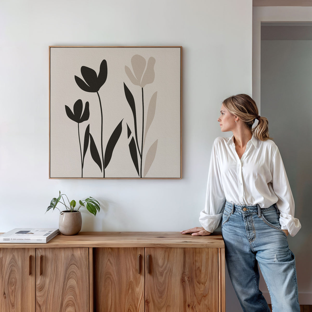 A woman in a white blouse and jeans leans against a wooden cabinet, gazing at the Floral Simplicity Canvas Art above her. A small potted plant and a book rest on the cabinet, enhancing the vintage-inspired decor of the scene.
