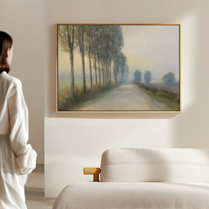 A woman stands in a softly lit room, gazing at the Forest Haze Canvas Art—a framed painting of a misty, tree-lined road. The space features a light-colored sofa and neutral décor.