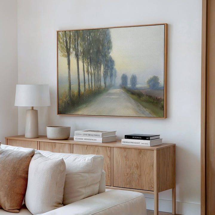 A cozy living room features a wooden sideboard with books, a table lamp, and decor. Above it hangs the Forest Haze Canvas Art, depicting a misty tree-lined road. A white couch is partially visible in the foreground.