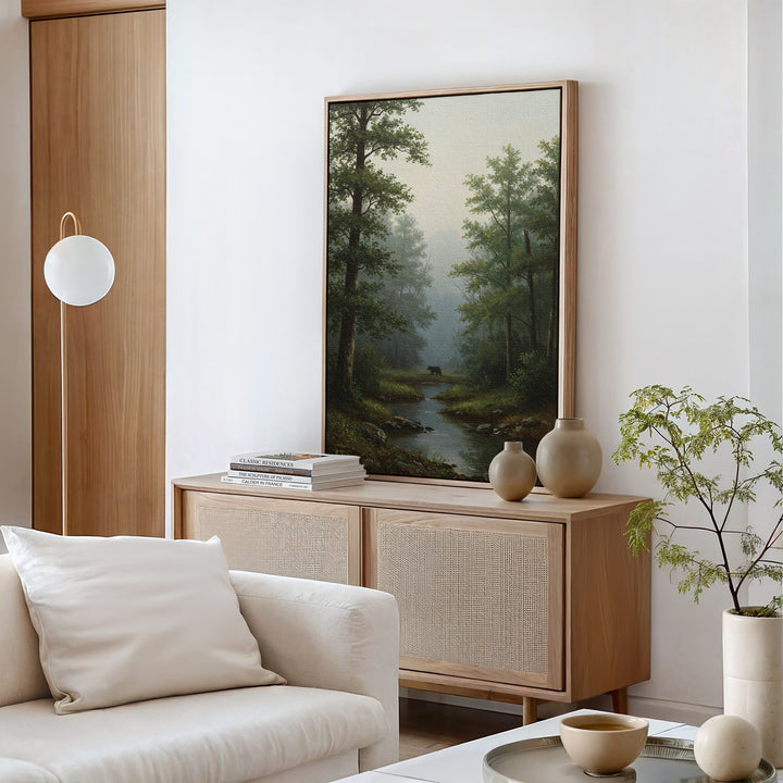 A modern living room featuring a light wood sideboard, neutral decor, the Forest Passage Canvas Art of trees and a stream, a round white lamp, a white sofa with cushions, and a potted plant.