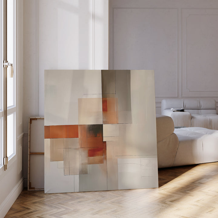 The Fractured Light Canvas Art, featuring warm beige, orange, and gray geometric shapes, rests on the floor of a modern living room with light walls, parquet flooring, and a white sofa.
