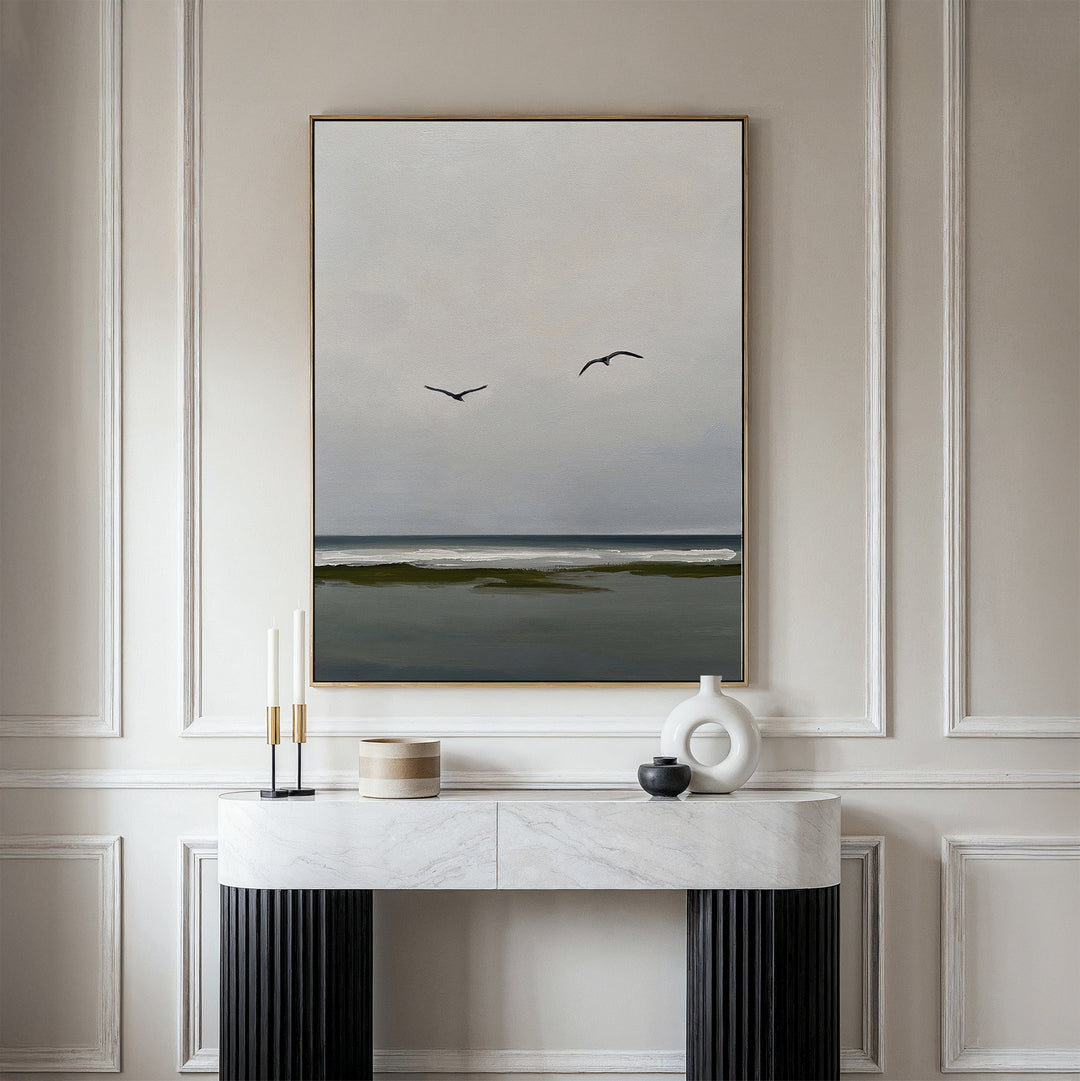A minimalist room features the Freeflight Canvas Art—a framed painting of a calm sea with two flying birds and a cloudy sky—above a white console table. Elegant molding and neutral tones amplify the serene, tranquil ambiance.