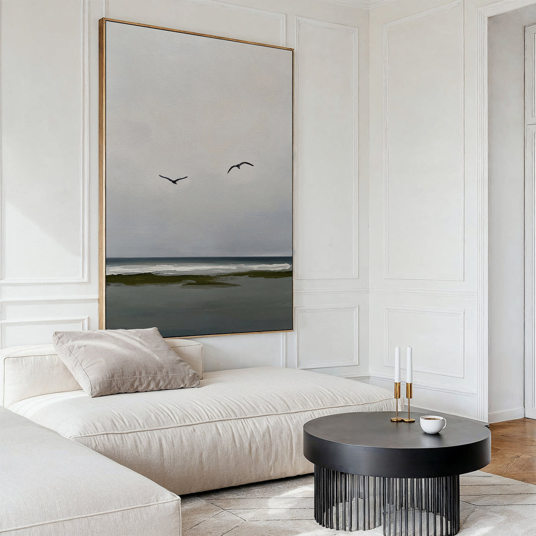 A modern, minimalist living room features a cream sofa, black round coffee table, candles, cup, and serene decor highlighted by the Freeflight Canvas Art—large wall art depicting an ocean view with two birds flying.